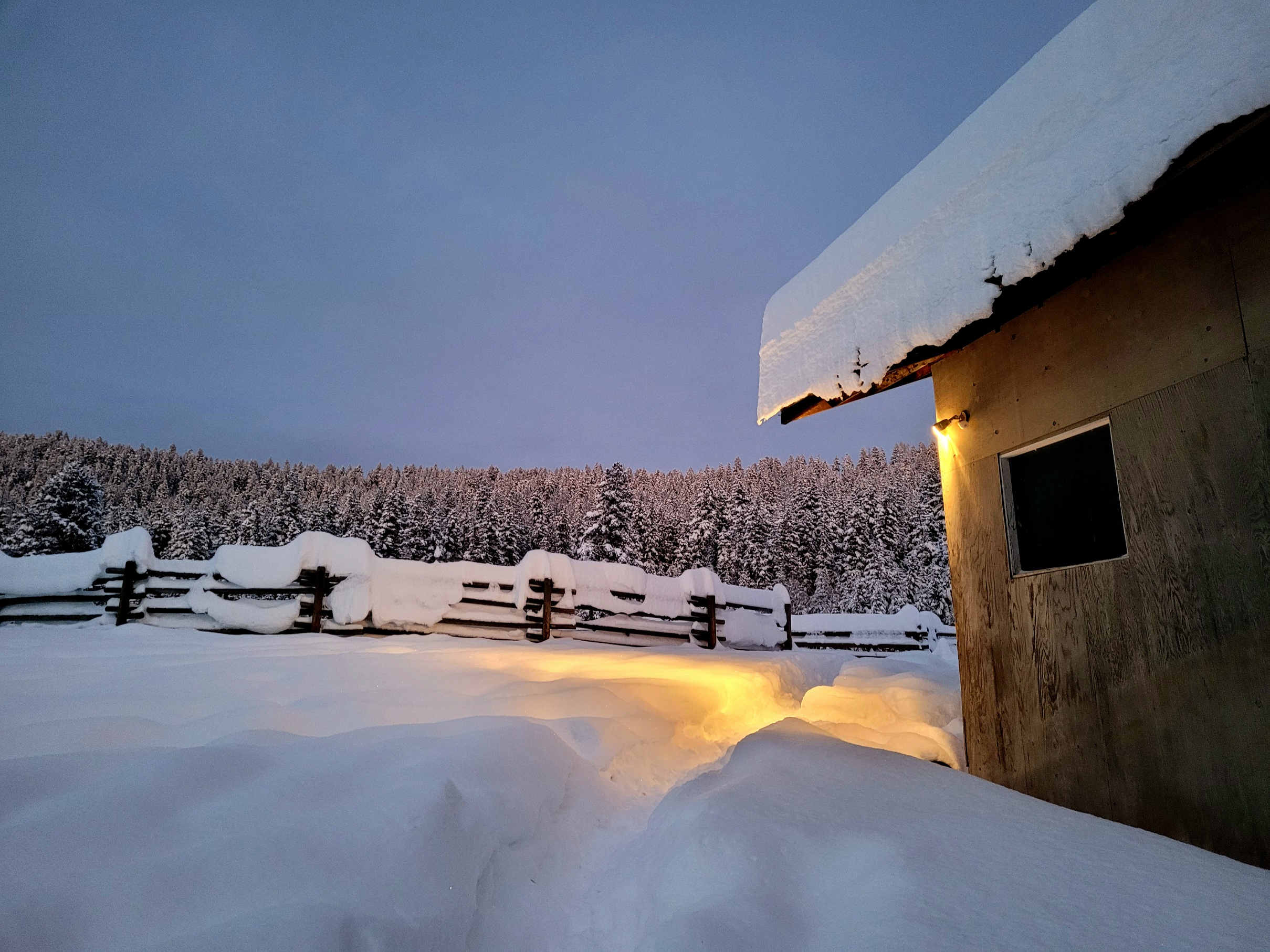 photo of the edge of a barn in heavy snow, getting close to dark, with a single light glowing on the snow