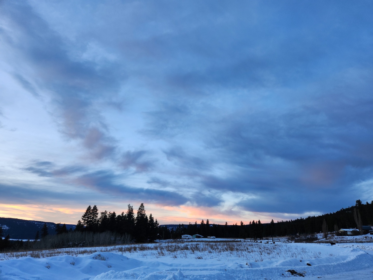 photo of a distance ranch across a snowy field, with sunset in a blue-grey low ceiling of clouds