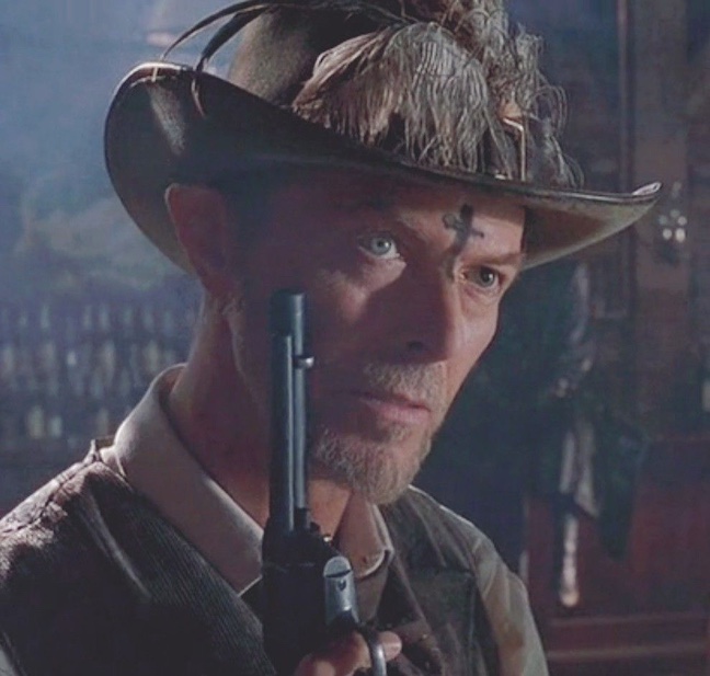 screenshot of David Bowie as Jack, holding up a pistol. a black upside down cross painted on his forehead
