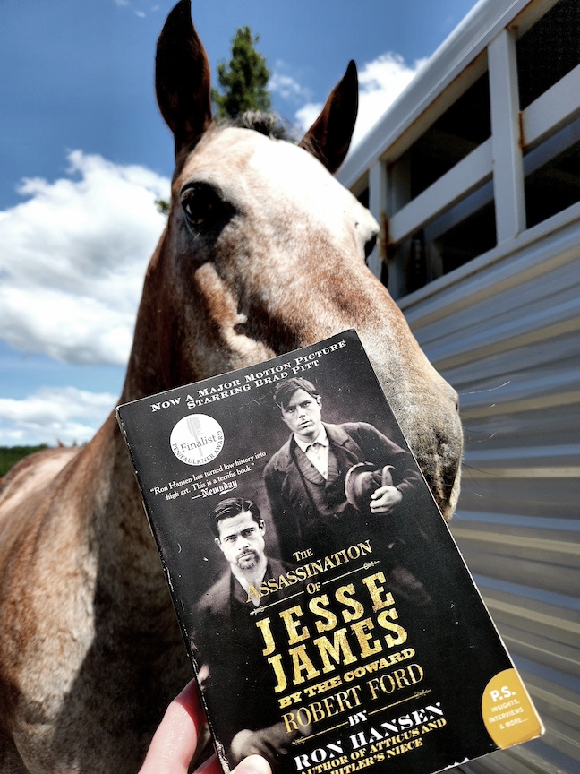 photo of the paperback The Assassination of Jesse James held up in front of an appaloosa horse standing beside a horse trailer