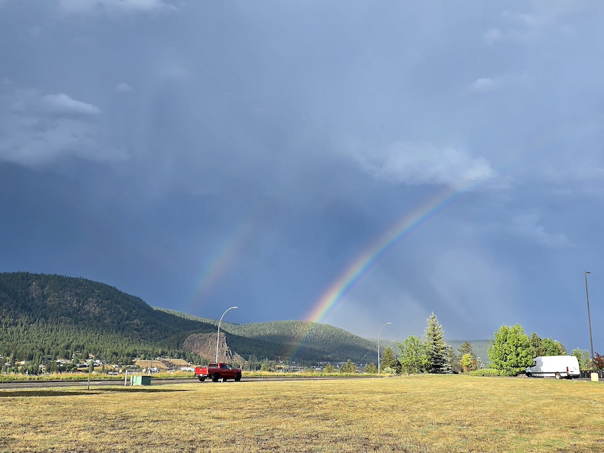 a double rainbow in a big dark blue sky, with a red pickup truck in the distance across yellow grass